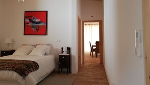 Una camera da letto con un letto, un comodino, una lampada, un quadro e vista su una sala da pranzo attraverso una porta aperta.