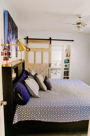 Un dormitorio con una cama, una mesita de noche, una lámpara, una estantería y una puerta que da al baño.