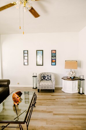 Una sala de estar con una mesa de centro de vidrio, un sillón estampado, un armario blanco y un ventilador de techo.