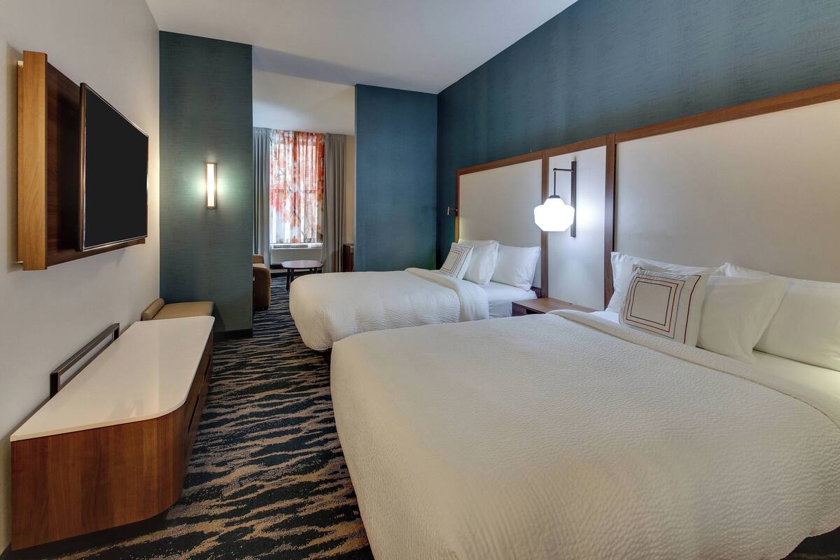 A hotel room with a large bed, a smaller bed, a desk with a chair, a flat-screen TV mounted on the wall, and a bathroom with a bathtub.