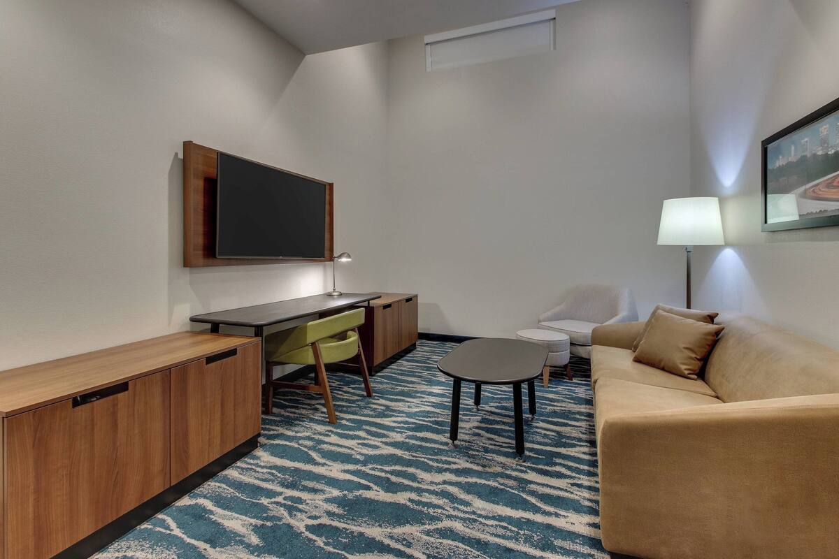 A compact hotel room with a wooden desk, a sofa, a small table, and a TV mounted on the wall.