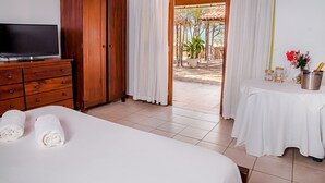 A hotel room with a bed, a TV on a wooden dresser, a table with a bottle of champagne, and a view of an outdoor patio with plants and a pergola. - Villa del Mar Praia hotel - Frente a Praia (Camocim)