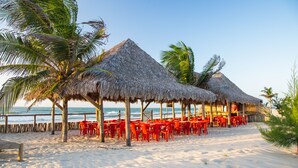 A beachside restaurant with thatched-roof structures, red tables and chairs, and palm trees. - Villa del Mar Praia hotel - Frente a Praia (Camocim)