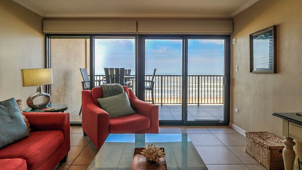Un salon moderne avec un canapé rouge, une table basse en verre et une vue sur la plage grâce à de grandes portes coulissantes.