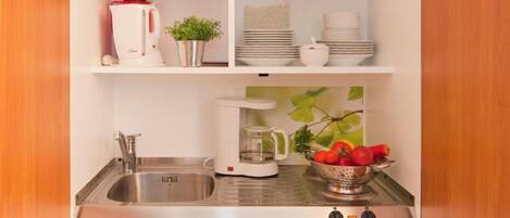 A compact kitchen with a sink, a coffee maker, a small refrigerator, and shelves with dishes and a plant.