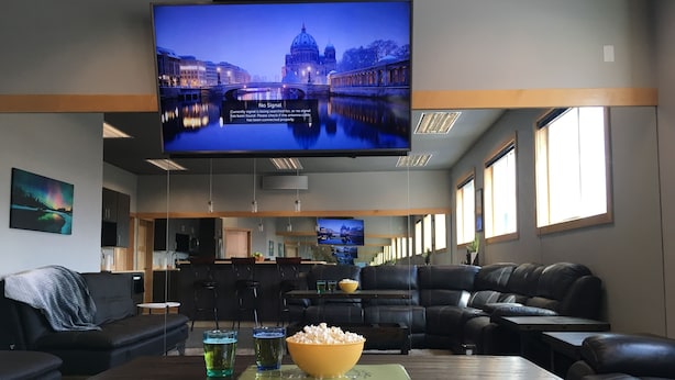 Een moderne woonkamer met een grote flatscreen televisie, een gezellige zithoek met leren banken, een eettafel met drankjes en snacks, en een keuken op de achtergrond.