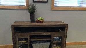 Een houten console tafel met een plant en een schaal appels erop. Het heeft open planken met boeken en een doos met de tekst 'Brain Games'.