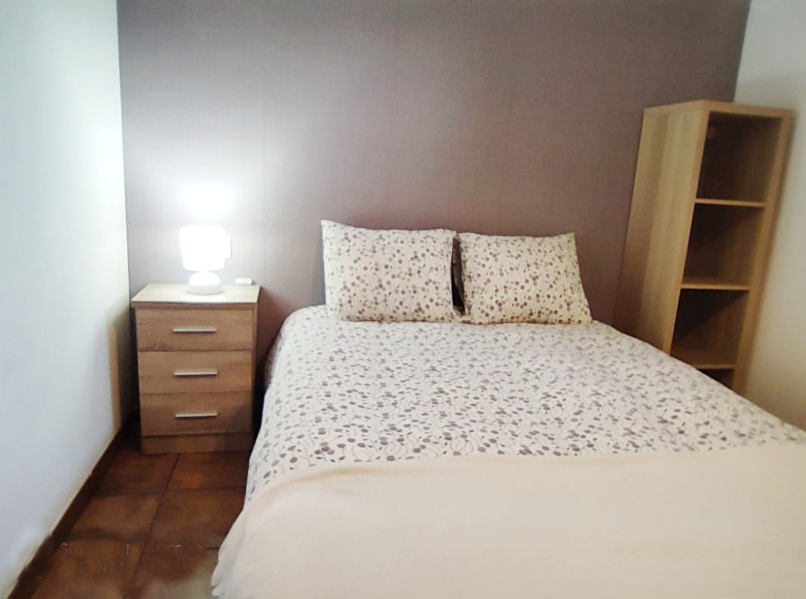 A bedroom with a bed, a nightstand with a lamp, and a wooden wardrobe.