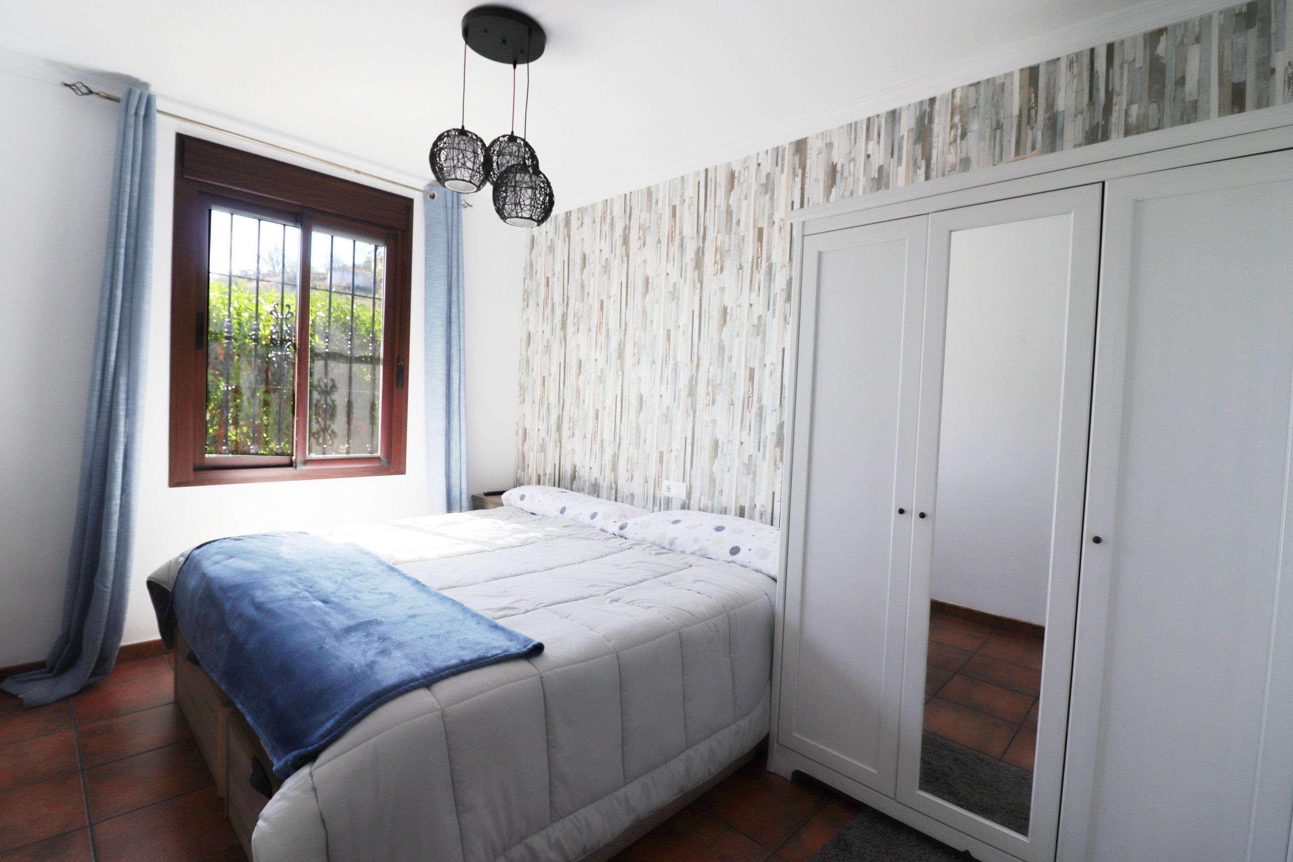 A bedroom with a bed, a wardrobe, a window with curtains, and a hanging light fixture.
