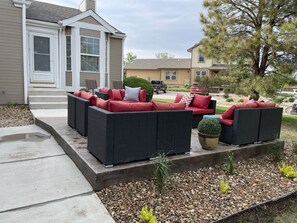 Eine Terrasse mit Korbmöbeln und roten Kissen, ein Betonweg und ein angelegter Garten mit Kies und kleinen Pflanzen.
