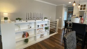 A modern living room with a white entertainment unit, wooden flooring, and a dining area with a black chair and pendant lights.