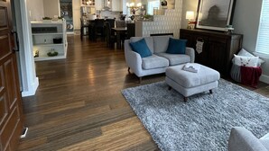 A spacious living room with a grey sofa, a coffee table, and a rug. There is a kitchen in the background with modern appliances and a dining area.