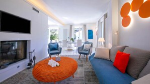 Ein modernes Wohnzimmer mit Flachbildfernseher, einem runden, orangefarbenen Hocker, einem blauen Sofa mit roten Kissen und einem Essbereich im Hintergrund.