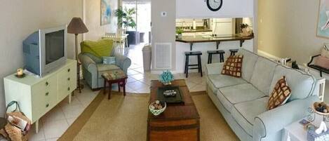 A living room with a sofa, a coffee table, a television, and a kitchen area in the background.