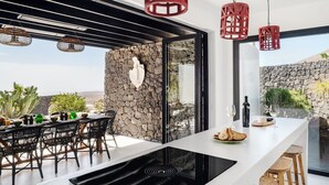 Una cocina moderna con pared de piedra, cocina por inducción negra y un comedor con sillas de mimbre y una mesa preparada para dos.