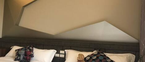Habitación de hotel con dos camas, cada una con ropa blanca y almohadas decorativas. La habitación presenta un diseño moderno con un patrón geométrico en el techo y un cabecero de madera oscura.