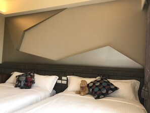Habitación de hotel con dos camas, cada una con ropa blanca y almohadas decorativas. La habitación presenta un diseño moderno con un patrón geométrico en el techo y un cabecero de madera oscura.