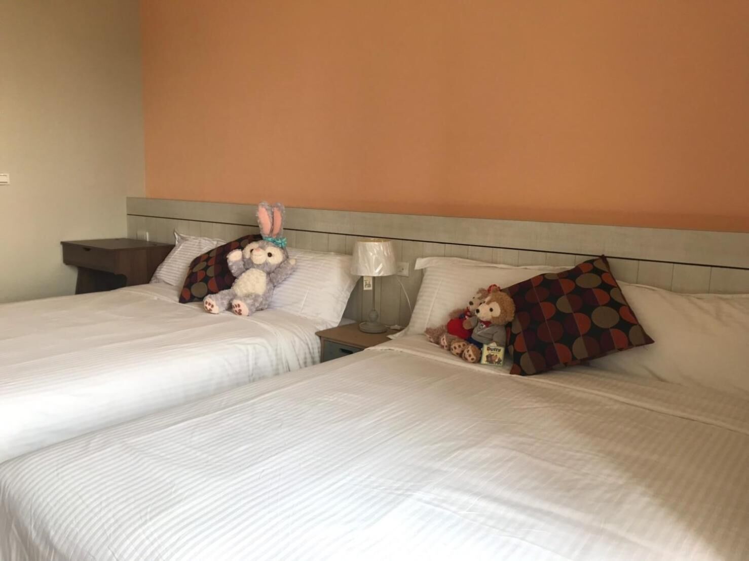 Een hotelkamer met twee bedden, elk voorzien van witte beddengoed en een hoofdbord. Twee nachtkastjes met lampen staan ernaast, en op een van de bedden ligt een teddybeer.