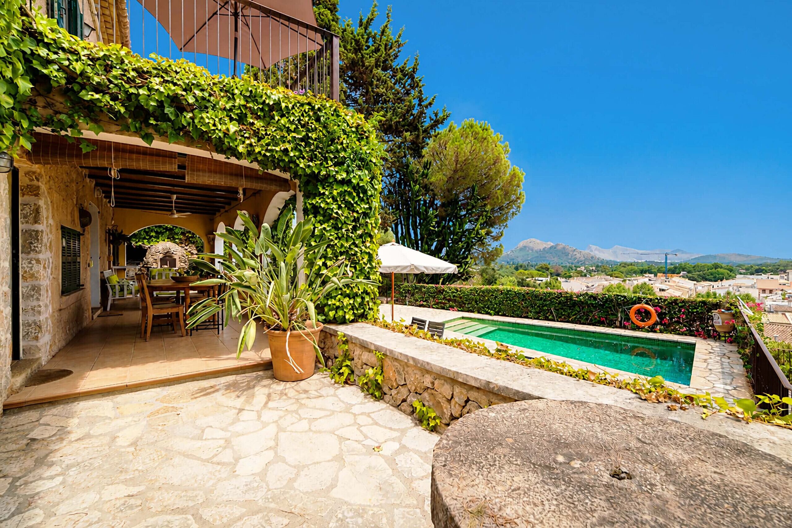A stone patio with a pool, a table with chairs, and a pergola covered with greenery.