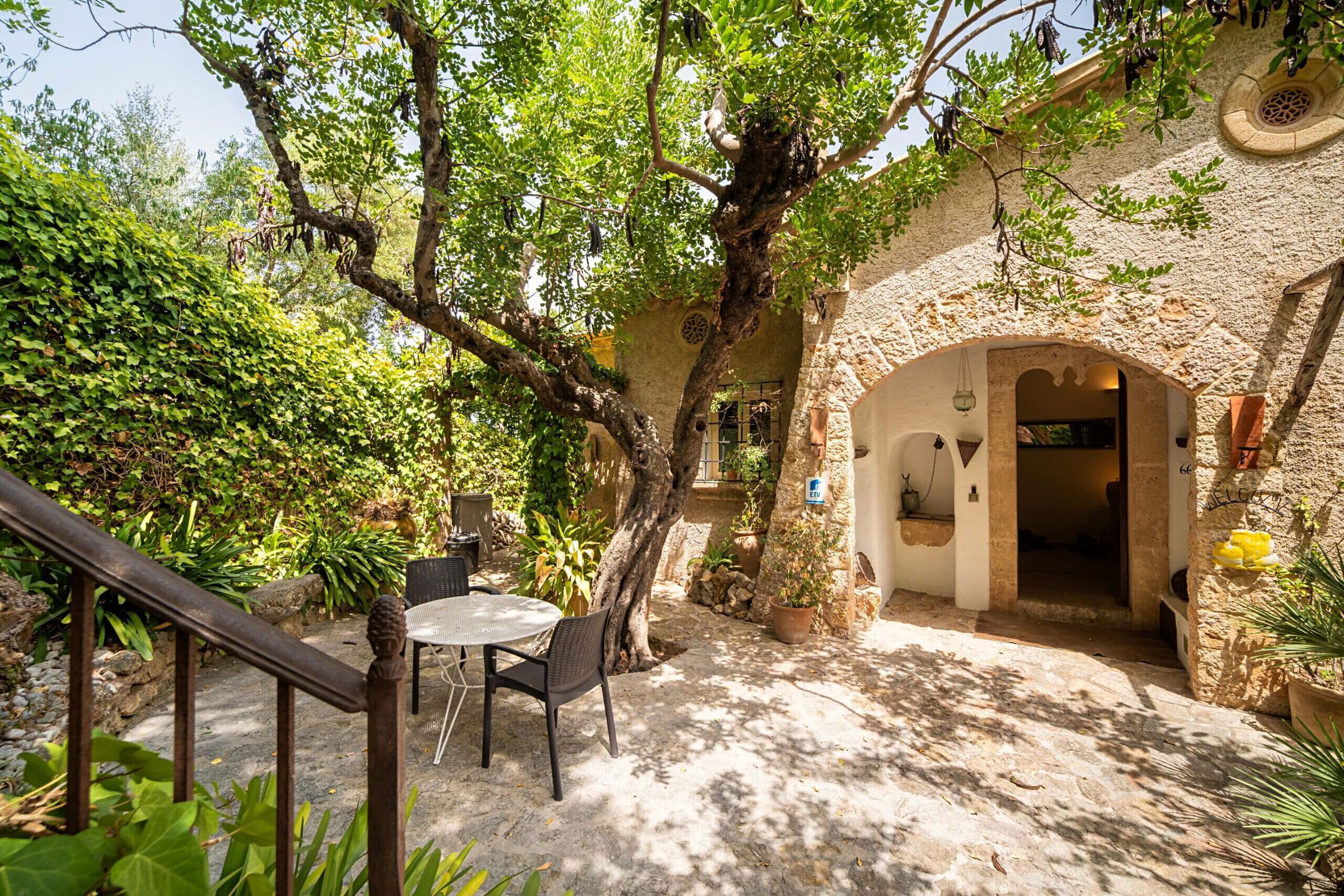 A courtyard with a table and chairs, surrounded by greenery and a stone building with an archway.