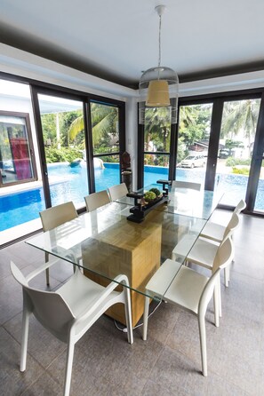 Ein moderner Essbereich mit einem Glastisch und weißen Stühlen, mit Blick auf einen Pool und Palmen.