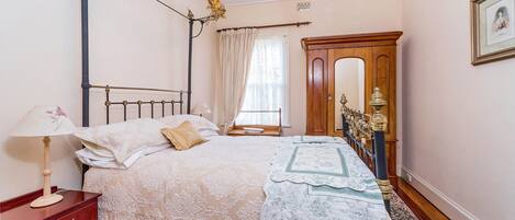 A bedroom with a four-poster bed, a wooden wardrobe, a nightstand, a lamp, and a framed picture on the wall.