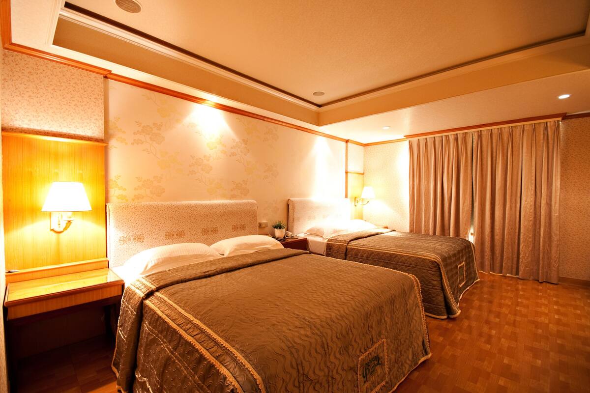 A hotel room with two beds, a nightstand with a lamp, a wall-mounted shelf, and a large window with curtains.