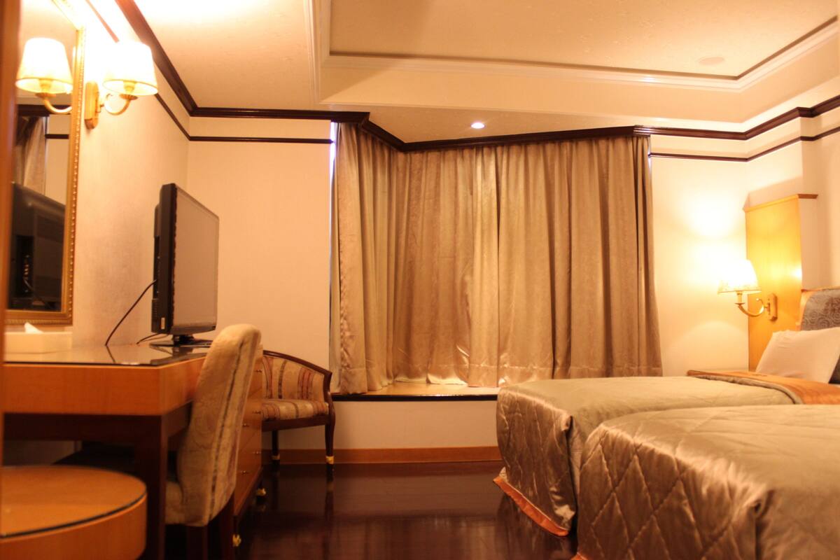 A hotel room with a bed, a desk with a chair, a television, and a window with curtains.