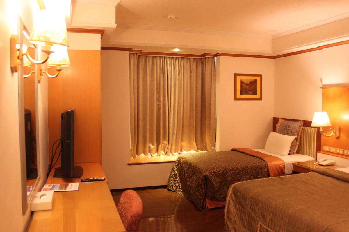 A hotel room with two beds, a desk with a computer, a TV, and a window with curtains.