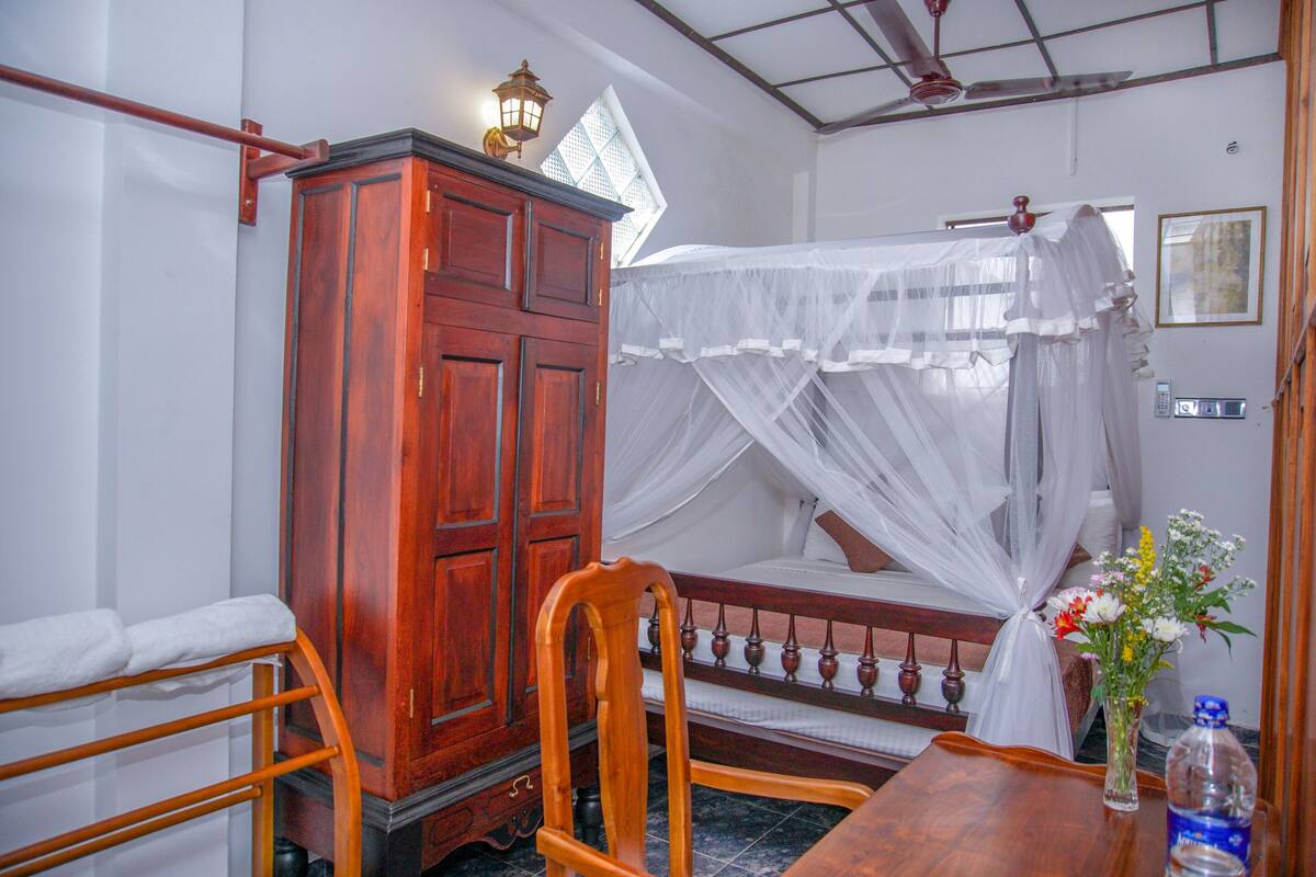 A room with a wooden cabinet, a bed with a canopy, a chair, a table, and a vase with flowers.