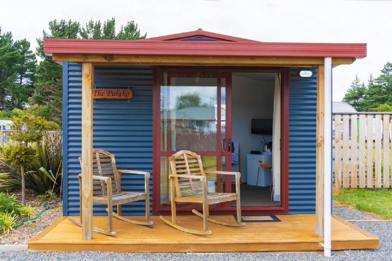 A small wooden structure with a red roof, labeled "The Pukeko," featuring two rocking chairs on a wooden platform.