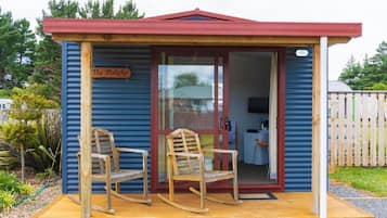 A small wooden structure with a red roof, labeled "The Pukeko," featuring two rocking chairs on a wooden platform.