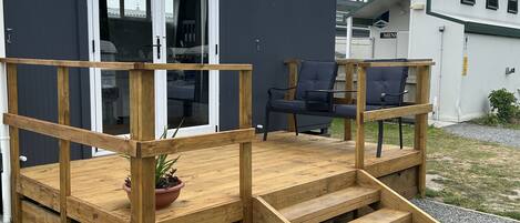 Una terraza de madera con una rampa que conduce a la entrada de un edificio, una planta en maceta y una silla al aire libre.
