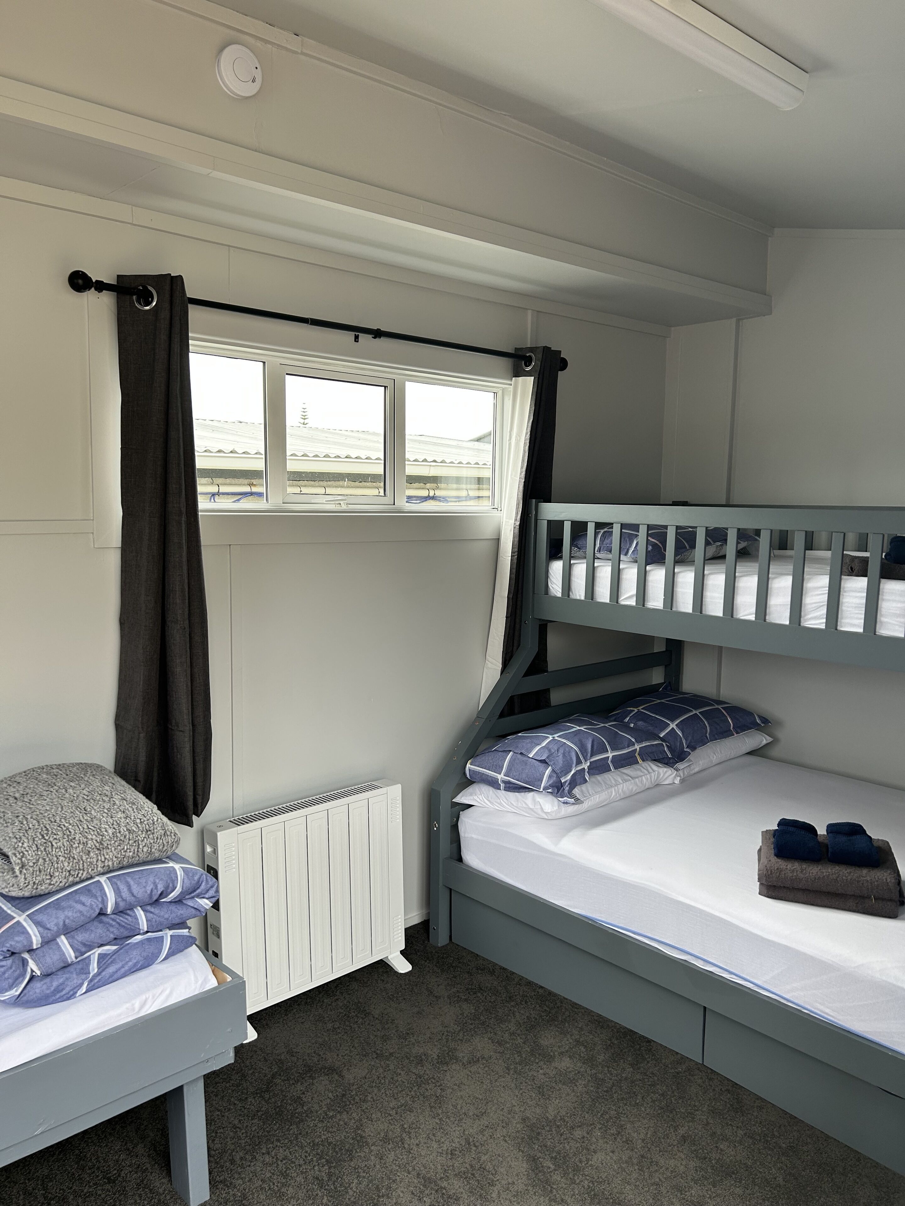 Een kleine kamer met stapelbedden, een raam met gordijnen, een radiator en een klein nachtkastje.