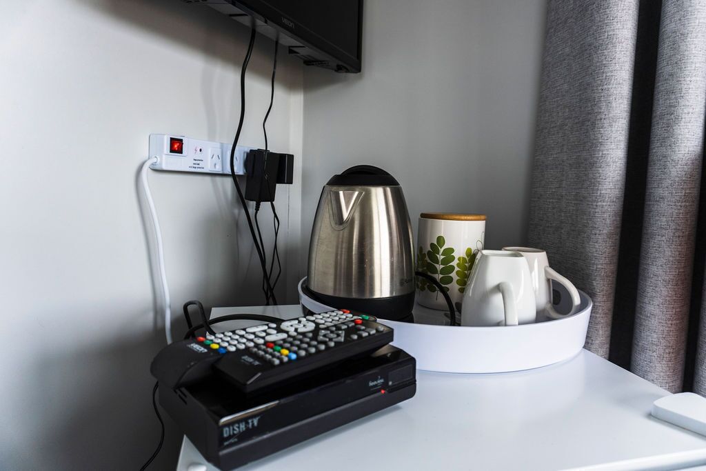 Een hotelkamer met een aan de muur bevestigde televisie, een koffiezetapparaat, een dienblad met mokken en een afstandsbediening op een tafel.