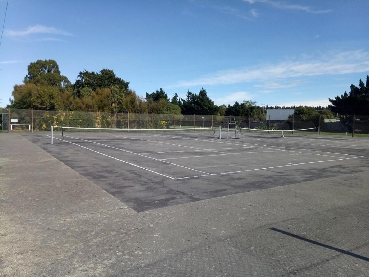 A tennis court with a net, surrounded by a fence and trees in the background.