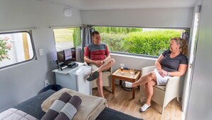 A compact RV interior with a bed, a small table, a TV, and a window offering a view of greenery. - Leithfield Beach Holiday Park (Leithfield)