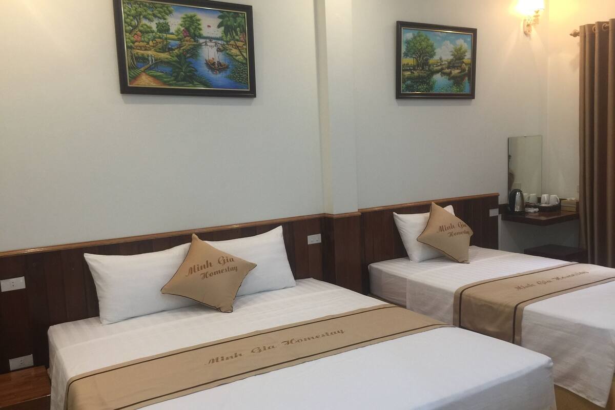 Two single beds with matching headboards and footboards, each with a pillow and a bed runner labeled "Minh Gia Homestay." There are two framed paintings above the beds, and a nightstand with a lamp to the right.