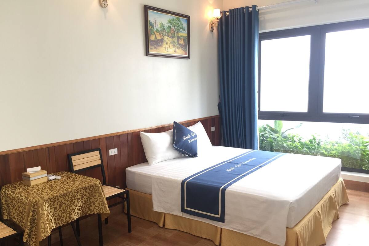 A hotel room with a bed, a table, and a chair. There is a window with curtains, a painting on the wall, and a view of greenery outside.