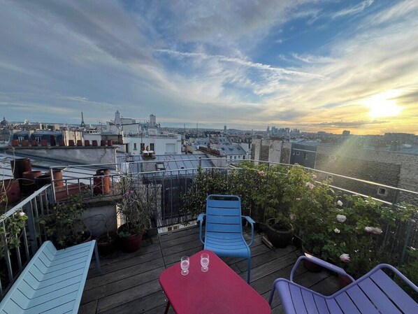 A rooftop terrace with a view of the city skyline, featuring a table and chairs, potted plants, and a railing.