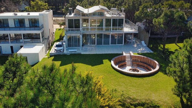 A modern multi-story house with a large deck, a circular outdoor seating area, and a view of a forested area.