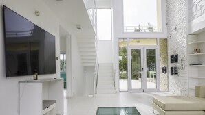 A modern living room with a flat-screen TV, a glass coffee table, a sofa, and a staircase leading to a balcony with glass doors.