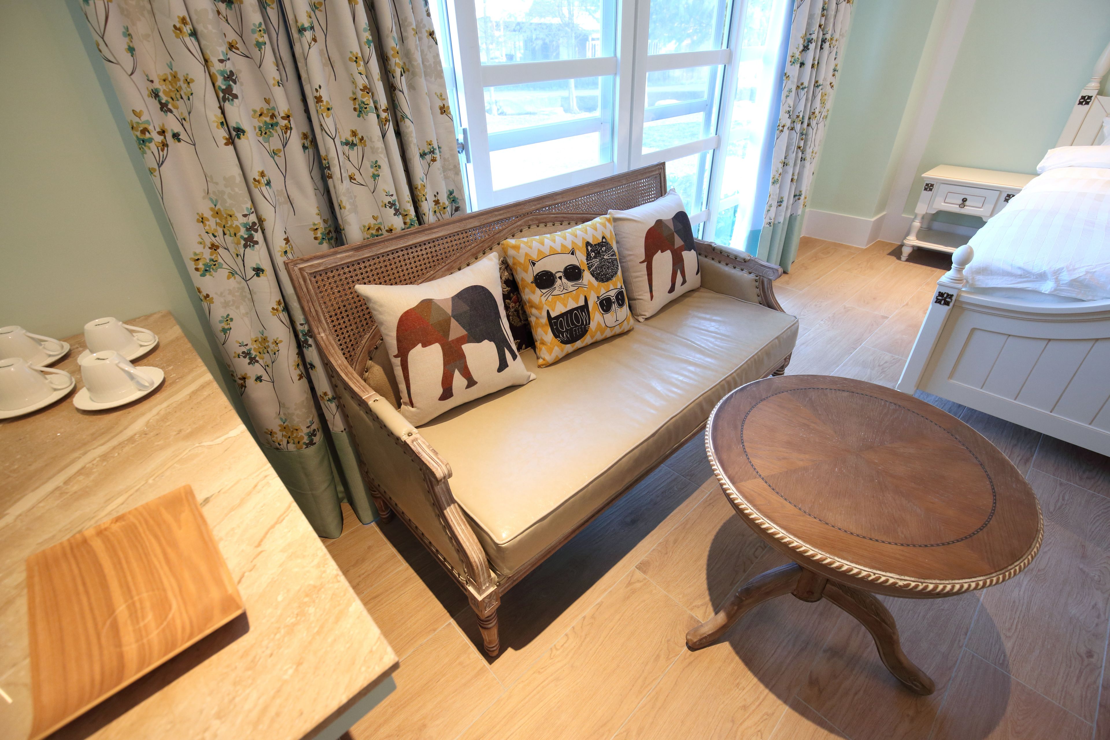 Een kamer met een slaapbank, een klein rond houten tafeltje en een raam met gordijnen.