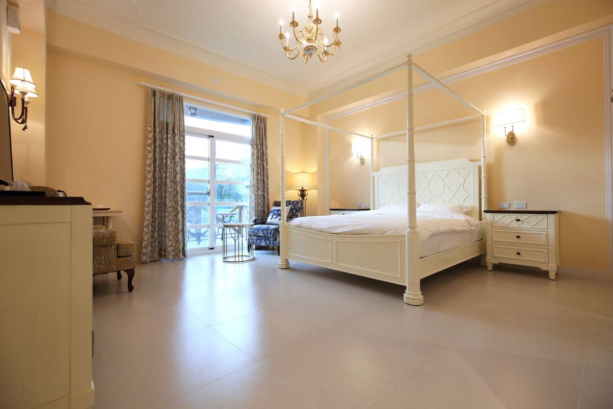 A four-poster bed with a nightstand, a chair, and a table in a well-lit room with a chandelier.