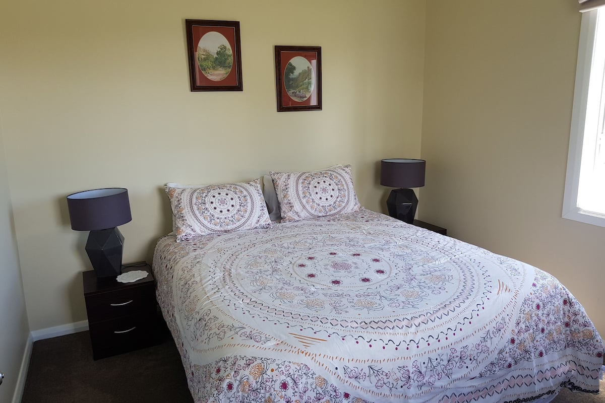 A bedroom with a large bed, two bedside lamps, a nightstand, and two framed pictures on the wall.