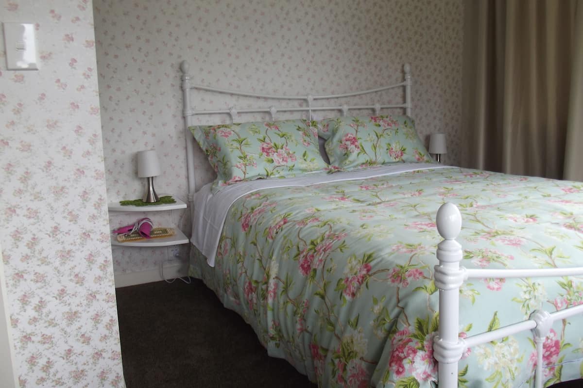 A neatly made bed with floral bedding, a bedside table with a lamp, and a wall-mounted light switch.