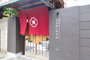 Entrada de hotel com uma cortina vermelha exibindo um logotipo branco, uma parede cinza com texto em japonês e uma pequena placa indicando "Room 1".