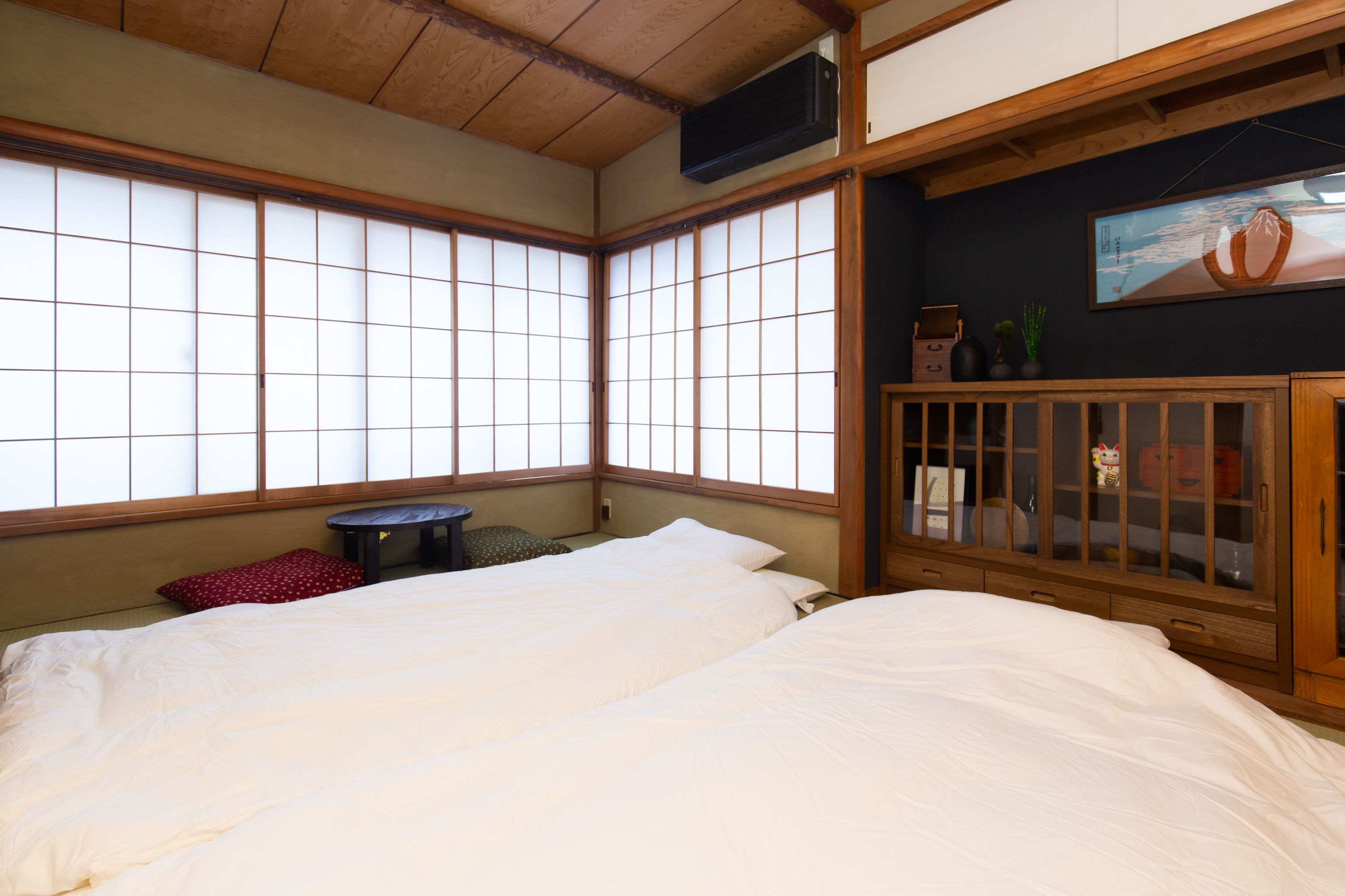 Una habitación tradicional japonesa con suelo de tatami, techo de madera bajo y puerta corredera tipo shoji. Hay una cama con ropa de cama blanca y una mesita con un cojín rojo.