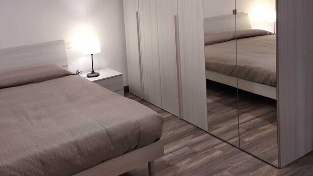 Een moderne slaapkamer met een bed, nachtkastje, lamp en een groot spiegelmeubel.