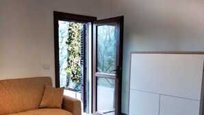 Een kamer met houten vloer, een bank, een kledingkast en een schuifdeur van glas die leidt naar een balkon met uitzicht op bomen.